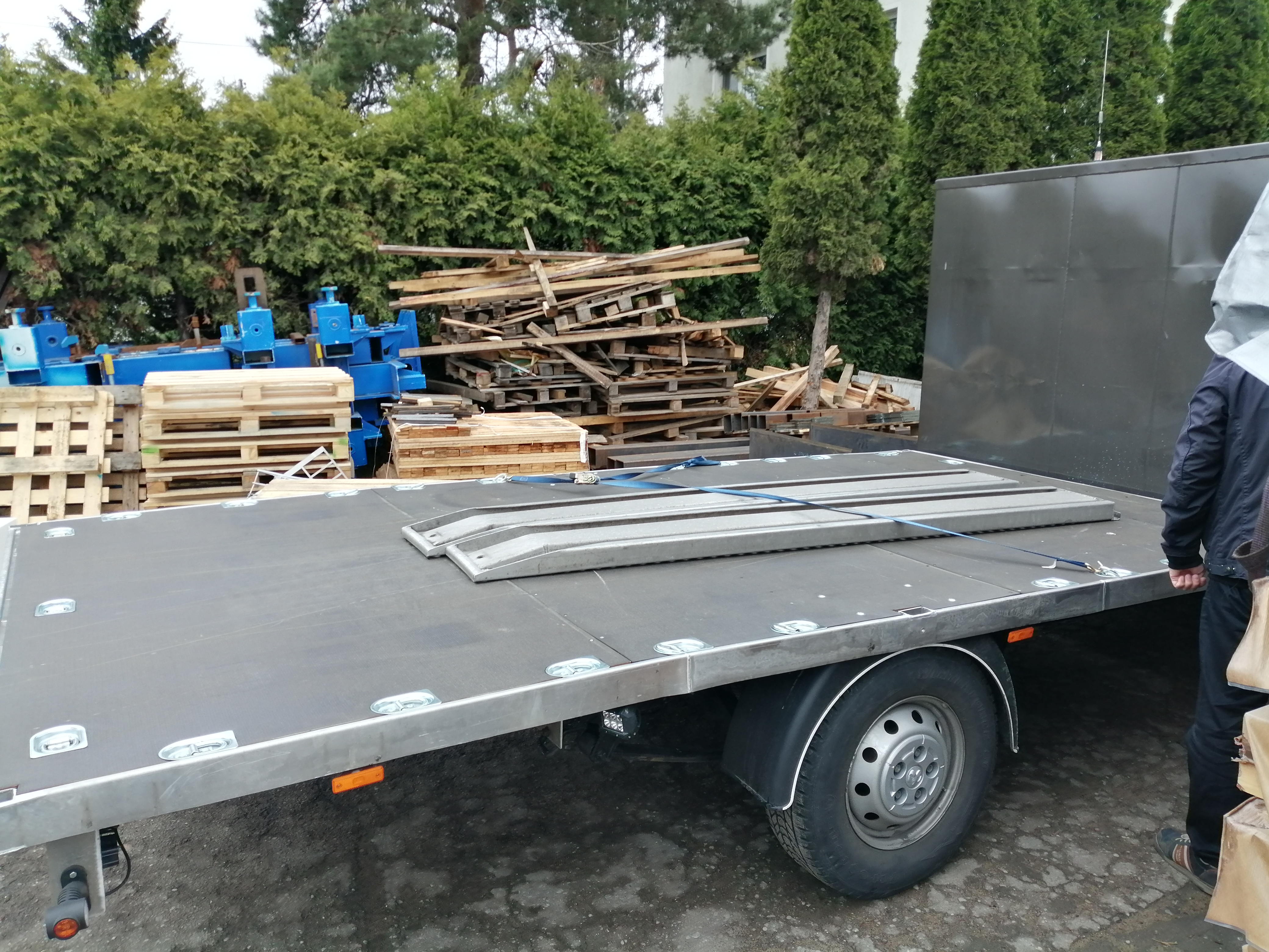 Przyczepa z platformą transportową, na której zabezpieczone są stalowe elementy konstrukcyjne, w tle drewniane palety i niebieskie elementy maszyn.