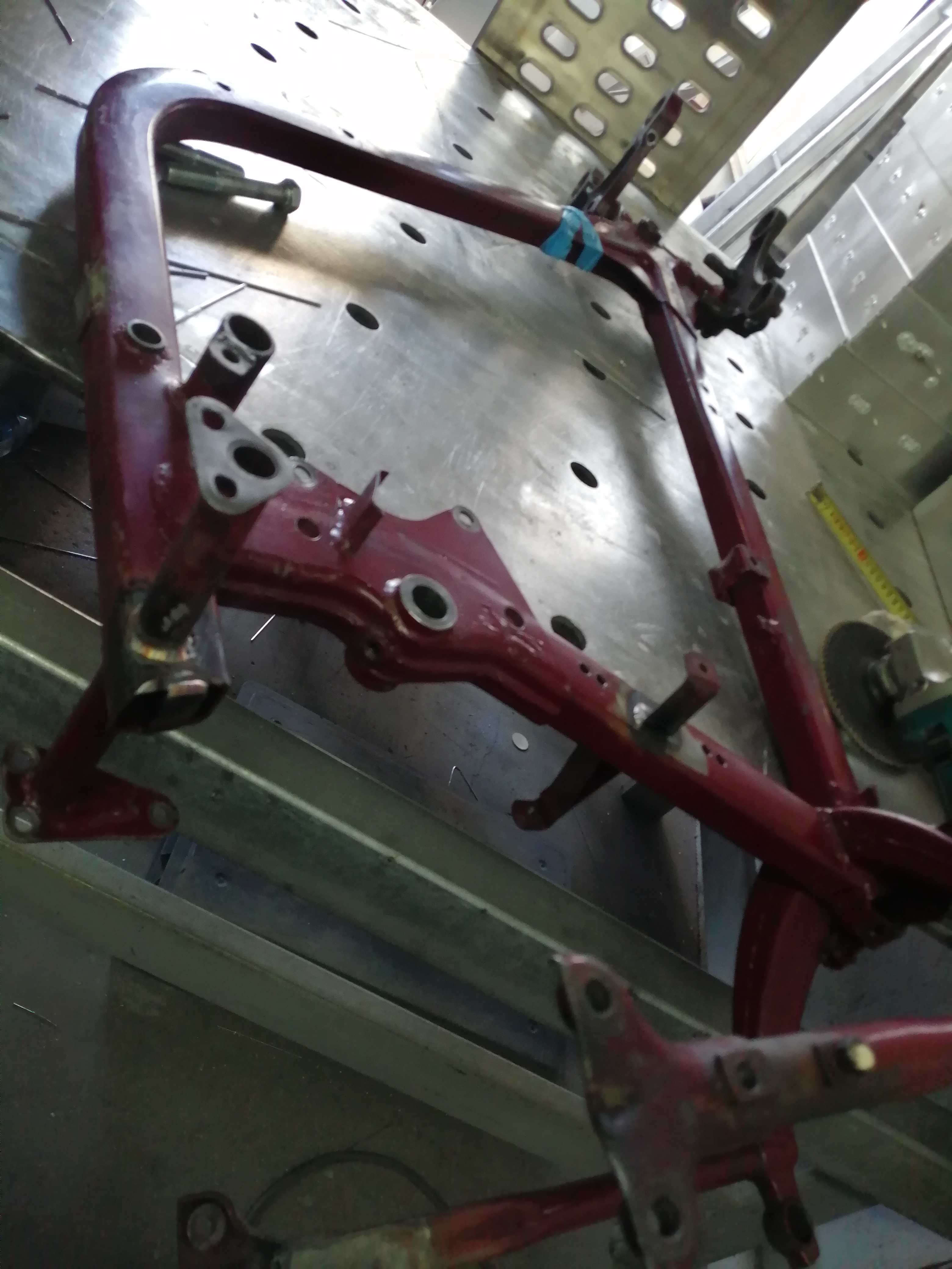 Czerwona, metalowa rama konstrukcyjna z licznymi otworami i elementami mocującymi, leżąca na perforowanym stole warsztatowym, prawdopodobnie w trakcie naprawy lub modyfikacji.