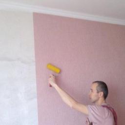 Wimaks - Mężczyzna maluje ścianę na różowo wałkiem z żółtym futrem, widoczna część ściany już pomalowana i fragment ściany w odcieniach bieli przed malowaniem.