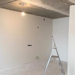 Wimaks - Świeżo pomalowane na biało wnętrze z widocznymi puszkami elektrycznymi i drabiną, surowy betonowy sufit z wiszącą żarówką.