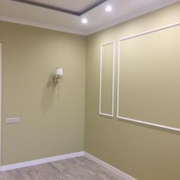 Wimaks - Pomalowane na oliwkowo ściany z białymi listwami przypodłogowymi i dekoracyjnymi ramkami, parkiet w odcieniach szarości, ozdobny kinkiet z abażurem.