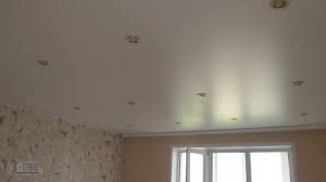 Biały, błyszczący sufit napinany z zamontowanymi punktowymi lampami halogenowymi, widok na fragment ściany z wzorzystą tapetą i okno.