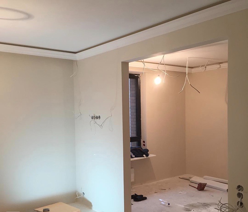 Pokój w trakcie remontu: ściany przygotowane pod malowanie, widoczne kable elektryczne, listwy sufitowe i okno z parapetem.