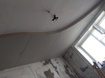 Niedokończone prace wykończeniowe wnętrza: surowy sufit z widocznym stelażem, ściany częściowo pokryte tynkiem, okno z parapetem, widoczna lampa sufitowa.