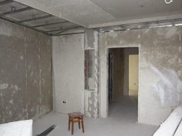 Surowe wnętrze w trakcie prac wykończeniowych: ściany pokryte szpachlą, widoczne profile sufitowe, mały stołek na środku pomieszczenia, niedokończone przejście do kolejnego pokoju.