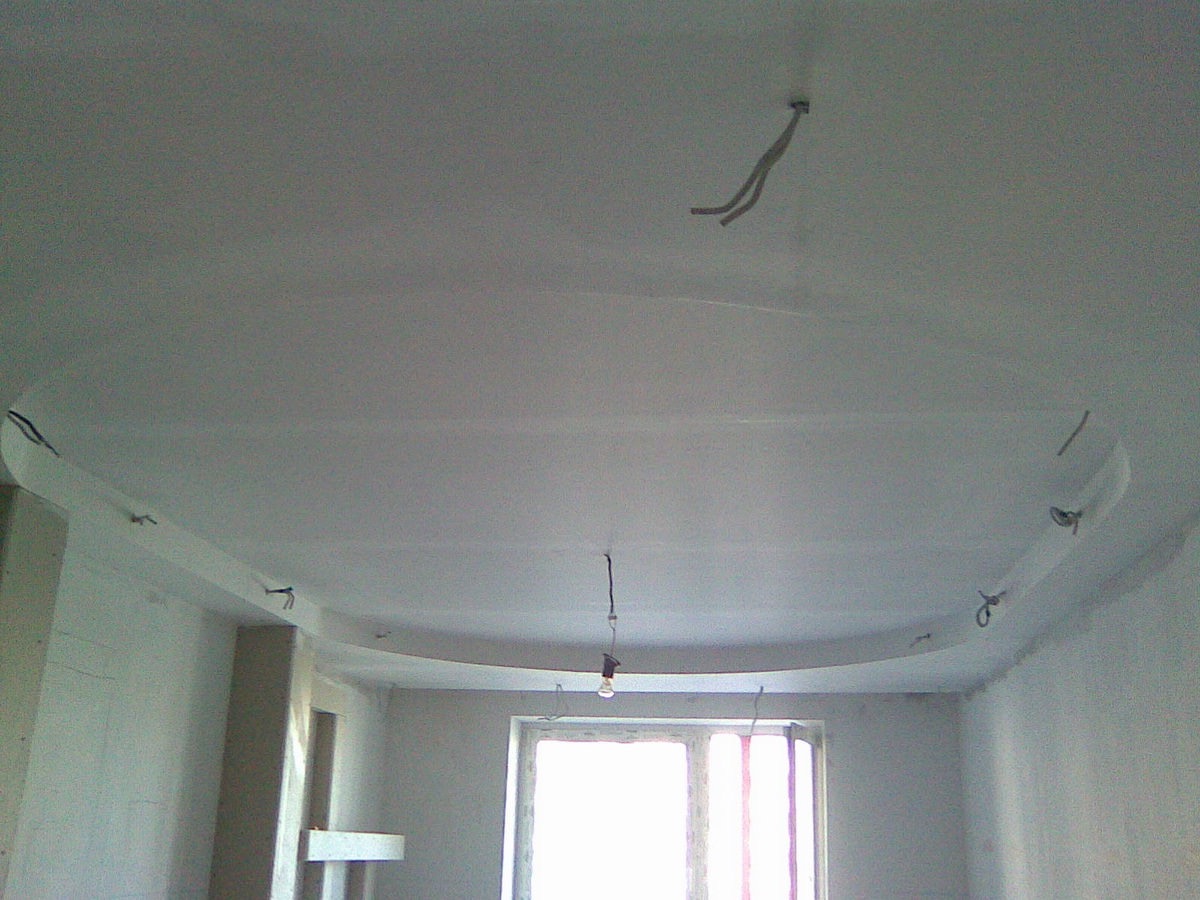 Niedokończony sufit podwieszany z wystającymi kablami i lampą wiszącą na tle okna w pokoju w stanie surowym.