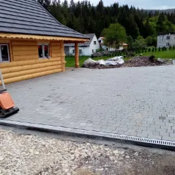 Ułożona szara kostka brukowa na posesji z drewnianym domem w stylu góralskim, widoczna maszyna do ubijania, krawężnik z odpływem liniowym oraz przygotowane podłoże z kamieni.