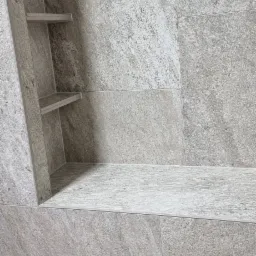 Narożna półka wnękowa z trzema półkami, wyłożona szarą płytką imitującą kamień, z odpływem liniowym w podłodze.