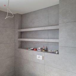 FIRMA BUDOWLANA RATAJCZYK EUGENIUSZ - Szara łazienka w trakcie wykończenia: płytki wielkoformatowe, wnęki z półkami, widoczne narzędzia i materiały budowlane, instalacja elektryczna sufitowa.