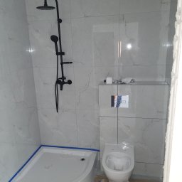 FIRMA BUDOWLANA RATAJCZYK EUGENIUSZ - Nowoczesna łazienka z kabiną prysznicową typu walk-in i czarną armaturą. Białe płytki imitujące marmur, podwieszana toaleta. Widoczne zabezpieczenia na brodziku.