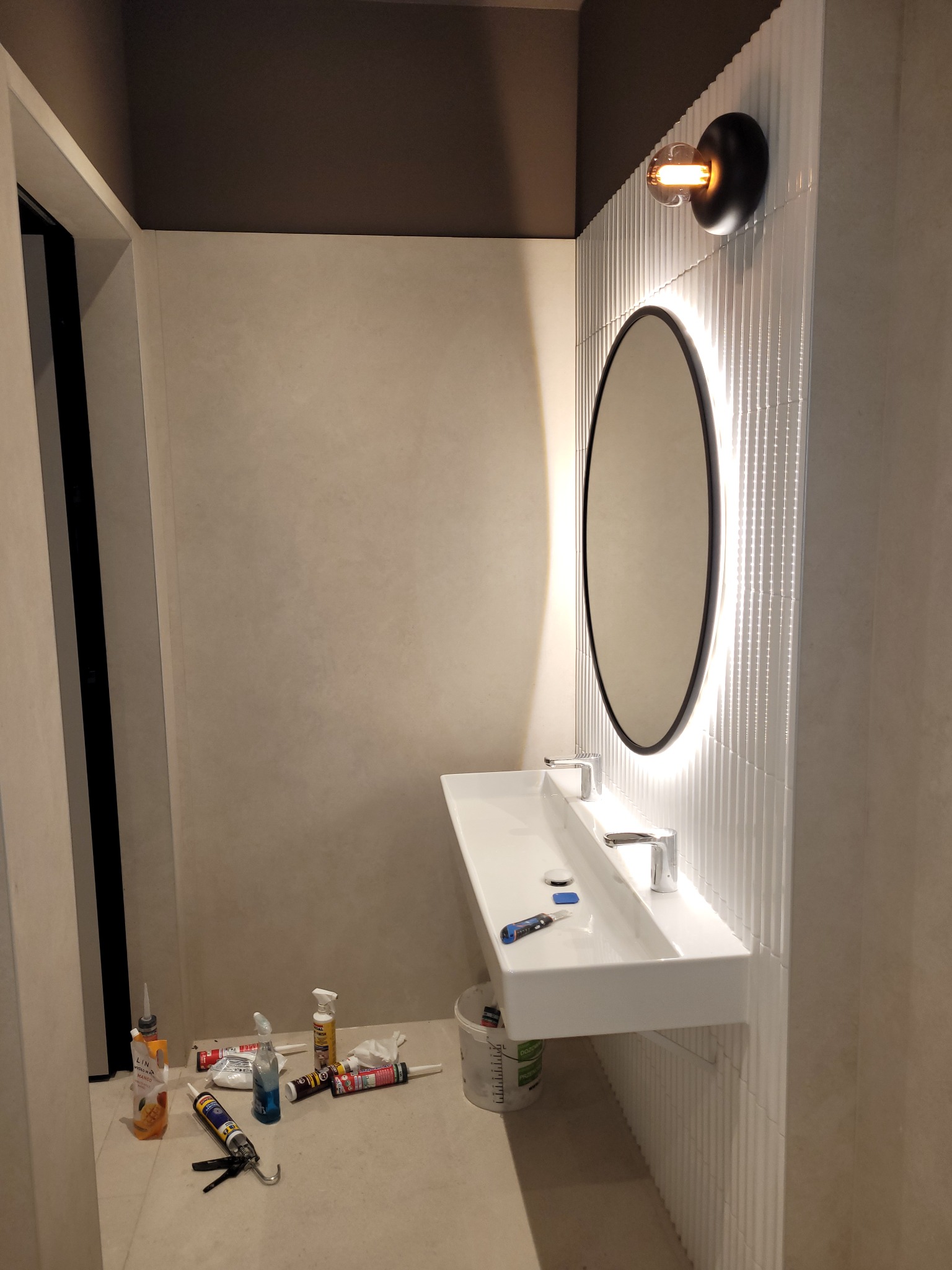 Nowoczesna łazienka w trakcie wykańczania: biała umywalka z armaturą, okrągłe lustro z podświetleniem LED i lampa w stylu retro na tle płytek z delikatnym wzorem.