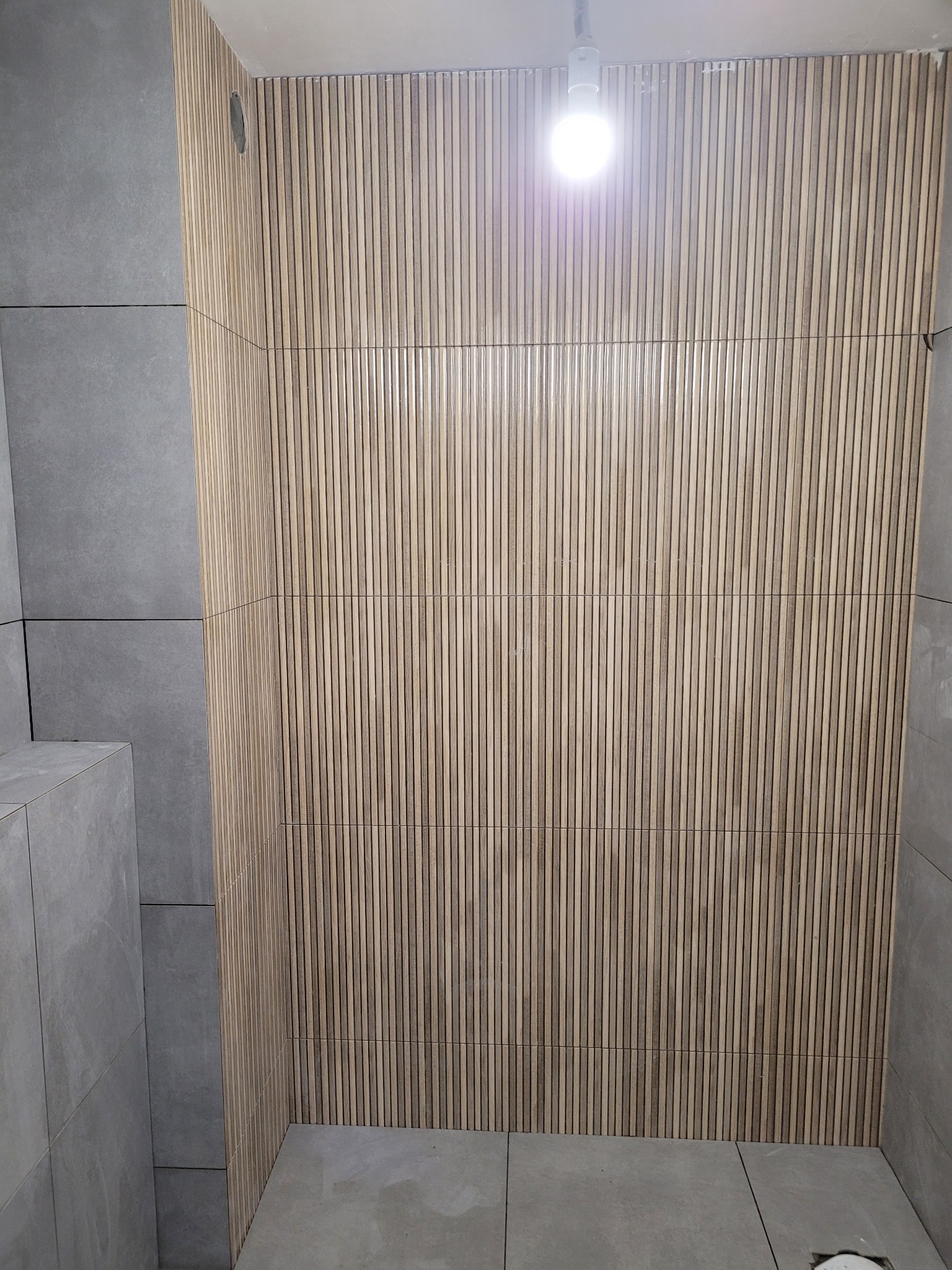 Wykończona ściana w łazience z drewnianymi lamelami i szarymi płytkami. Nowoczesny design, widoczne łączenia paneli i płytek. Oświetlenie sufitowe.
