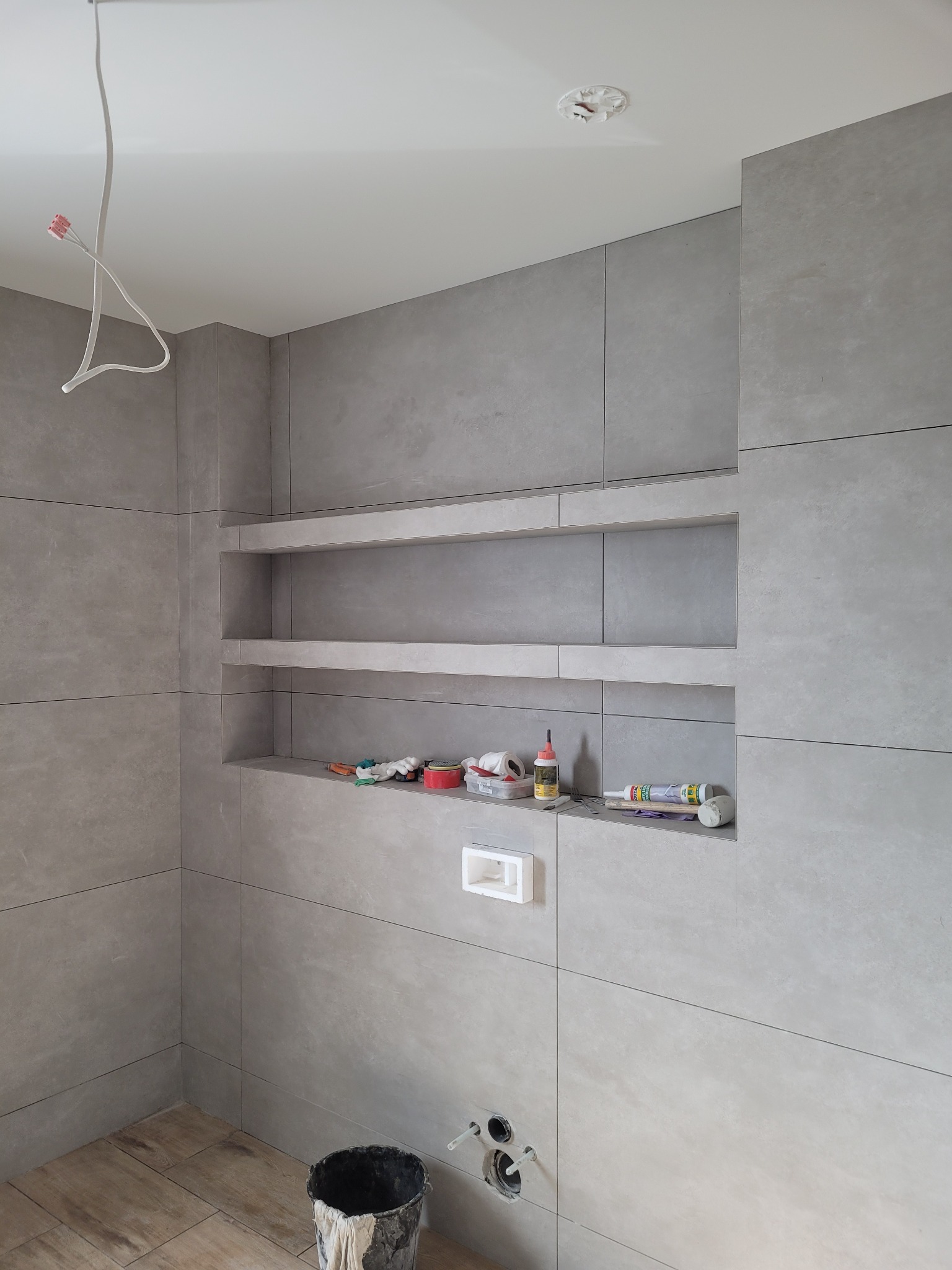 Szara łazienka w trakcie wykończenia: płytki wielkoformatowe, wnęki z półkami, widoczne narzędzia i materiały budowlane, instalacja elektryczna sufitowa.