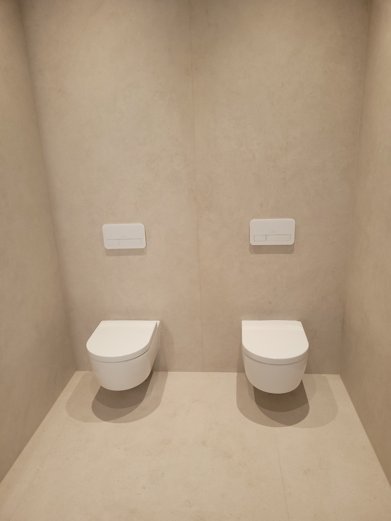 Dwa podwieszane białe sedesy z przyciskami spłukującymi na beżowej ścianie i podłodze z płytek imitujących kamień w wykończonej łazience. Minimalistyczny design.