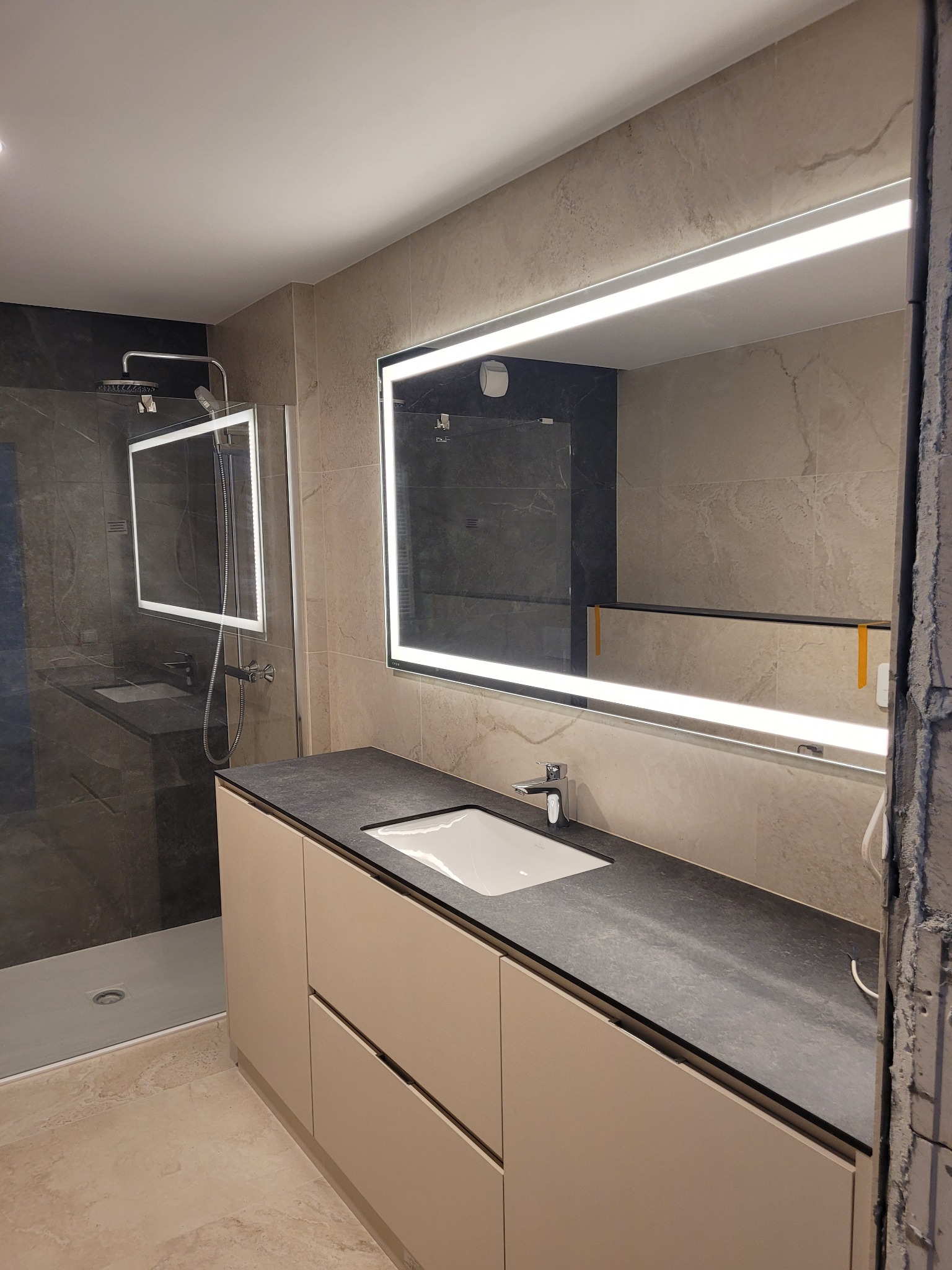 Nowoczesna łazienka po remoncie: beżowe płytki, podświetlane lustro, szafka z ciemnym blatem, kabina prysznicowa z ciemnymi płytkami. Widoczny fragment ściany w trakcie wykończenia.