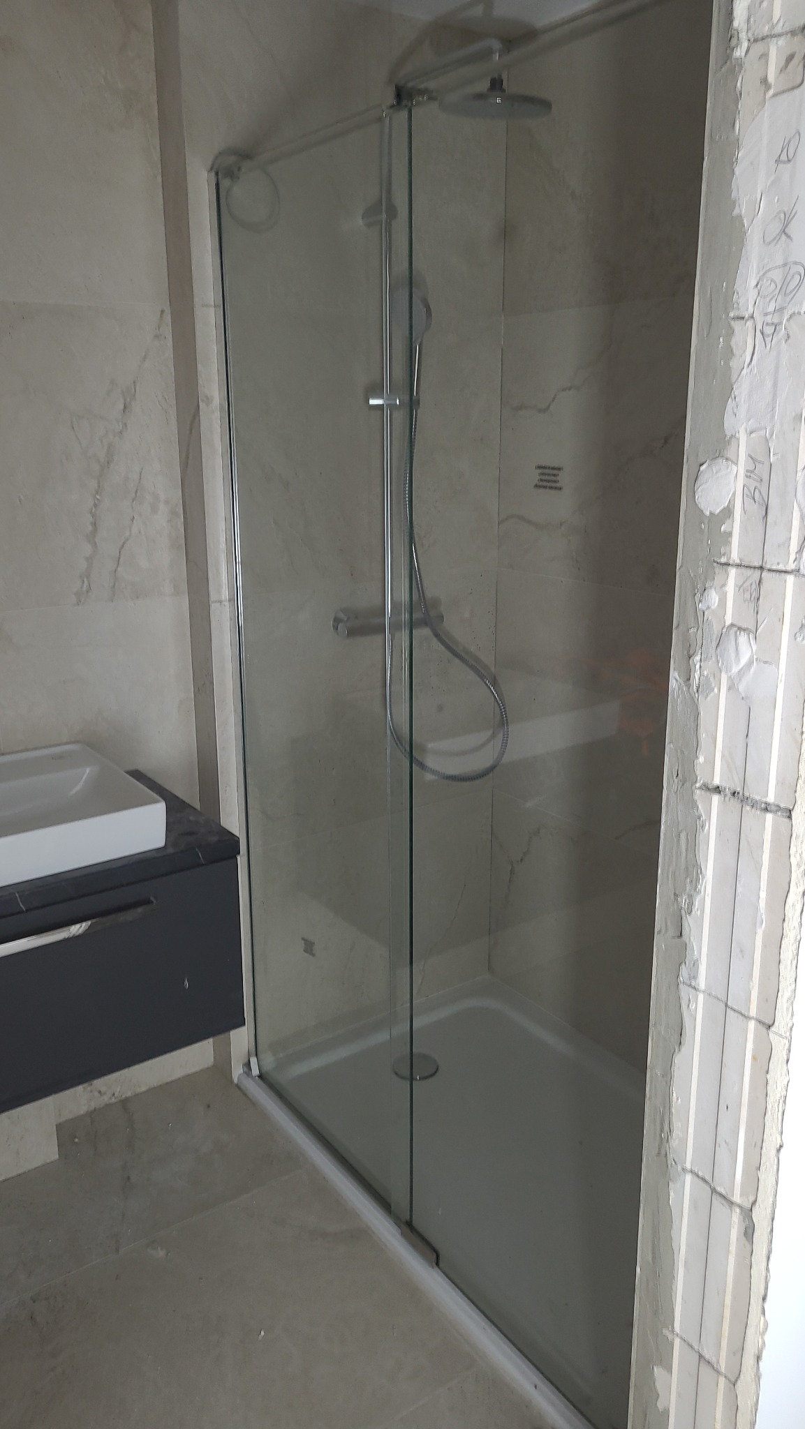 Kabina prysznicowa ze szklanymi drzwiami, marmurowe płytki na ścianach, fragment umywalki i surowa ściana w trakcie prac wykończeniowych w łazience.