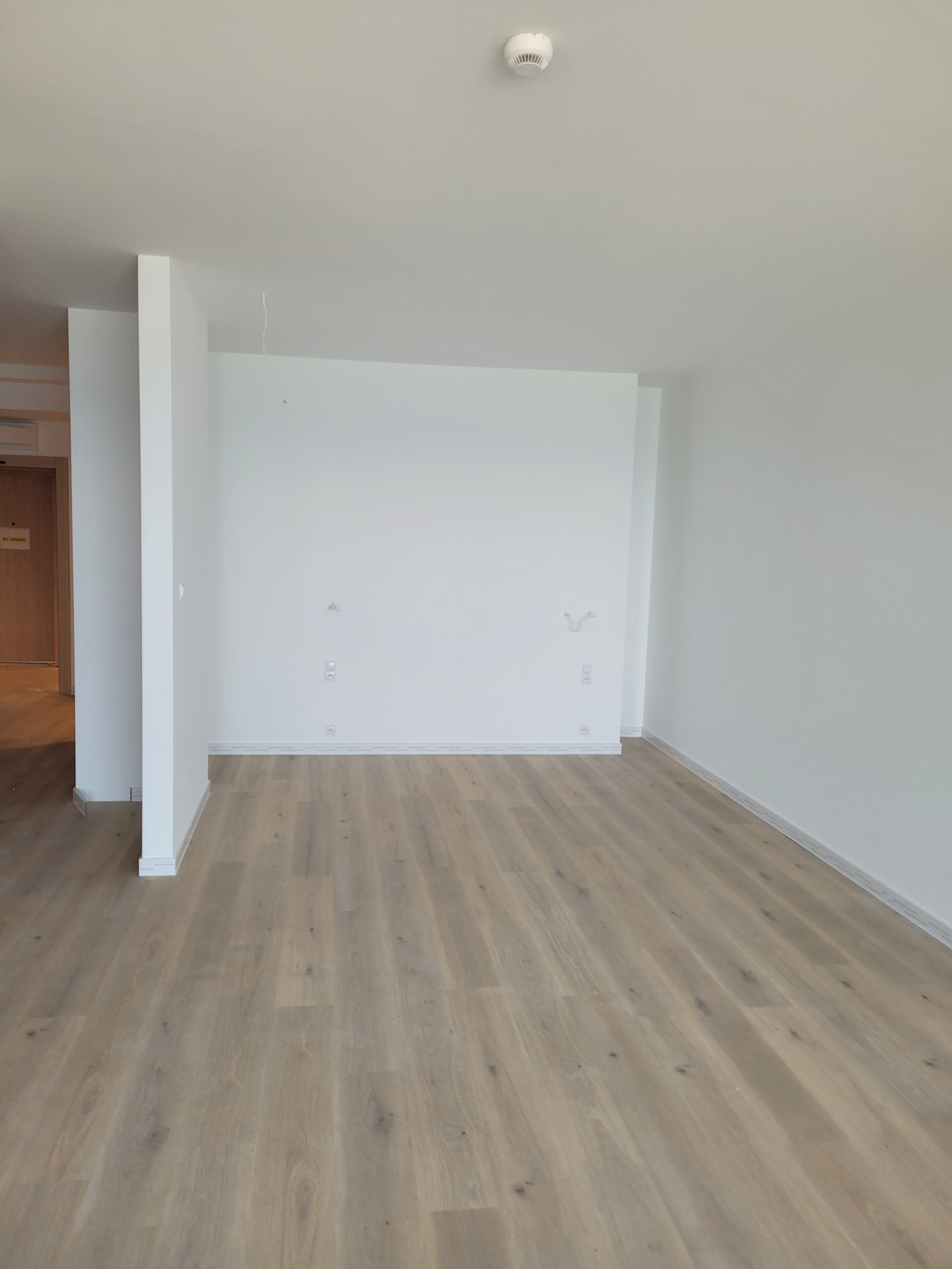 Jasne panele podłogowe w pustym, odmalowanym na biało pokoju z widocznymi gniazdkami i wyprowadzeniami kabli. Minimalistyczne wnętrze w stanie deweloperskim.