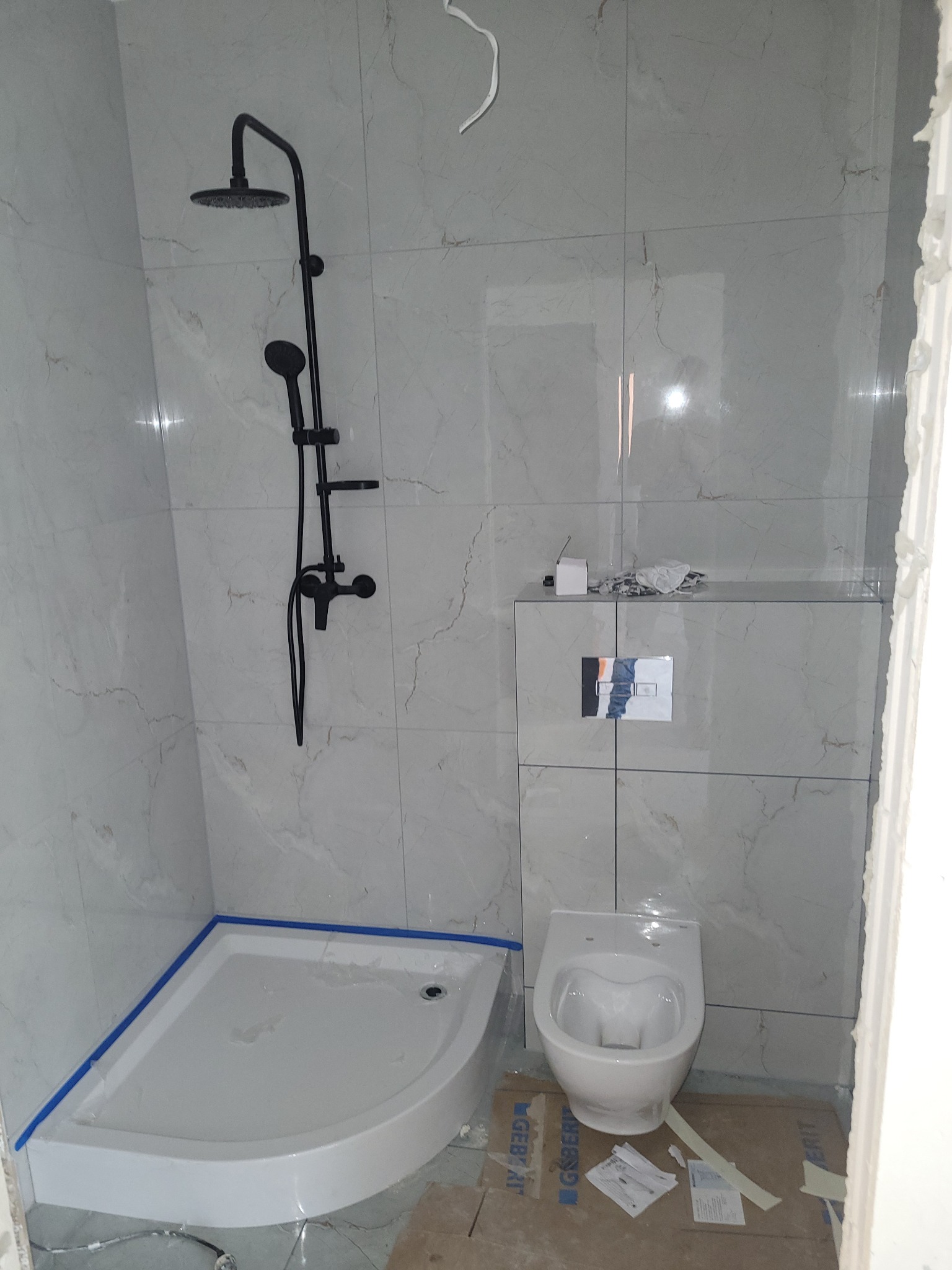 Nowoczesna łazienka z kabiną prysznicową typu walk-in i czarną armaturą. Białe płytki imitujące marmur, podwieszana toaleta. Widoczne zabezpieczenia na brodziku.