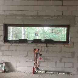 Surowa ściana z betonu komórkowego z poziomym oknem, widoczne ślady po pracach murarskich, instalacja wodno-kanalizacyjna zabezpieczona pianką montażową, napis 'KUCHNIA OKNO' na ścianie, w tle las.