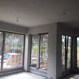 Surowy stan wnętrza z dużymi oknami przed wykończeniem, widoczne gładzie na ścianach i suficie, instalacja elektryczna wystająca z sufitu, na zewnątrz widok na las i materiały budowlane.