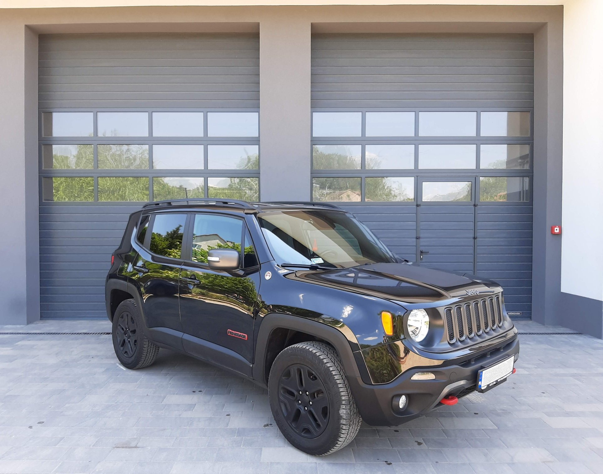 Czarny Jeep Renegade stoi przed garażem z dwoma szarymi bramami segmentowymi z oknami, widoczne odbicia drzew w szybach.