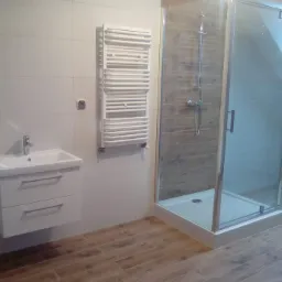 Nowoczesna łazienka z kabiną prysznicową, umywalką z szafką i grzejnikiem drabinkowym, wykończona płytkami imitującymi drewno na podłodze i jasnymi płytkami na ścianach, oświetlona naturalnym...