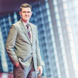 Portret mężczyzny w szarym garniturze i wzorzystym krawacie, z ręką w kieszeni, na rozmytym tle biurowca z dużymi oknami.