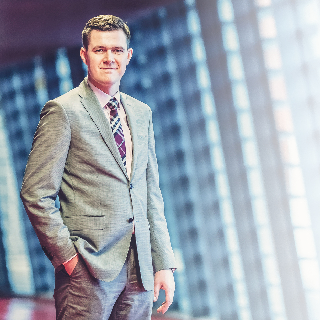 Portret mężczyzny w szarym garniturze i wzorzystym krawacie, z ręką w kieszeni, na rozmytym tle biurowca z dużymi oknami.