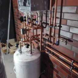Nowy bojler owinięty folią ochronną, podłączony do miedzianych rur grzewczych, zamontowany na ceglanej ścianie. Widoczne zawory i elementy instalacji w trakcie montażu.