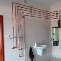 Miedziane rury instalacji wodnej zamocowane do ściany w łazience z umywalką i centralnym odkurzaczem, widoczne otwory w ścianie.