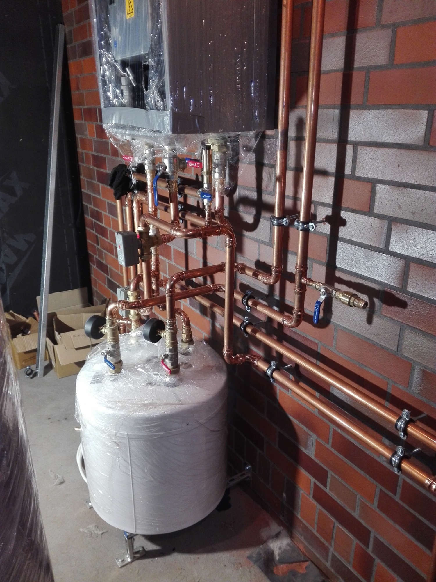 Nowy bojler owinięty folią ochronną, podłączony do miedzianych rur grzewczych, zamontowany na ceglanej ścianie. Widoczne zawory i elementy instalacji w trakcie montażu.