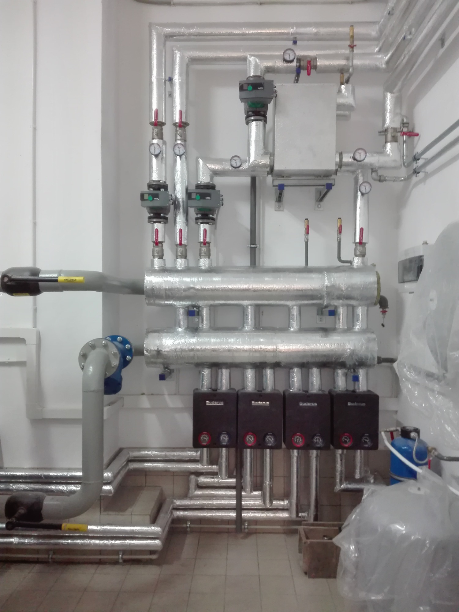 Rozbudowana instalacja grzewcza z izolowanymi rurami, zaworami i manometrami, czterema piecami Buderus oraz niebieskim filtrem wody, widoczna na tle białej ściany i podłogi z płytek.