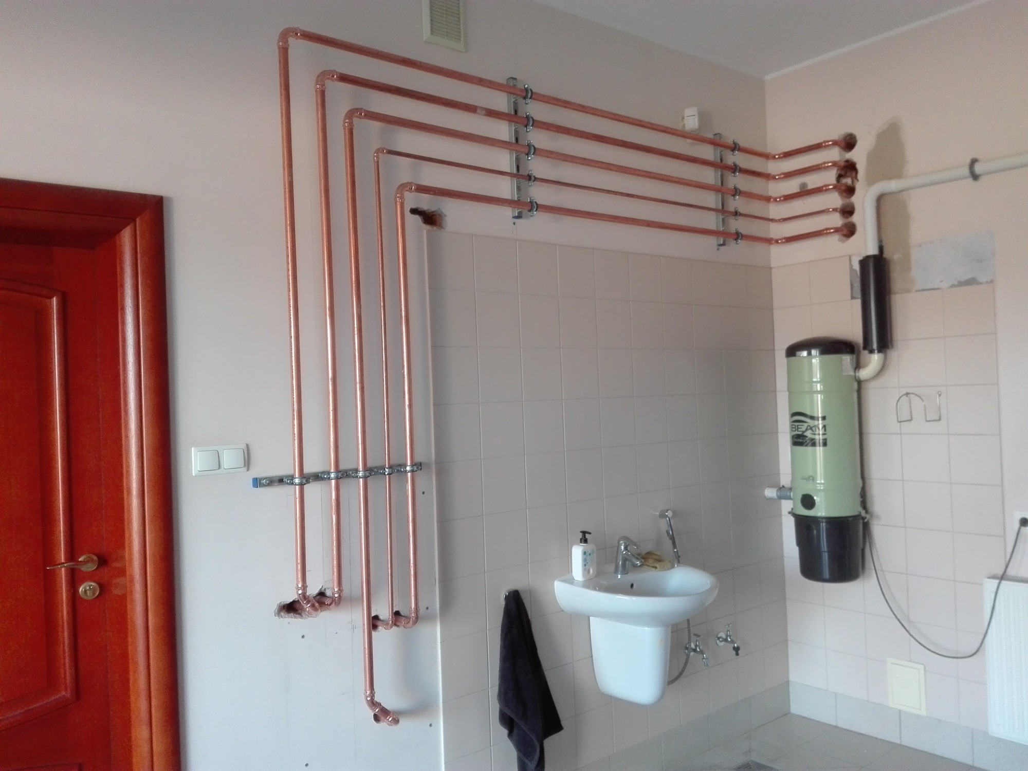 Miedziane rury instalacji wodnej zamocowane do ściany w łazience z umywalką i centralnym odkurzaczem, widoczne otwory w ścianie.