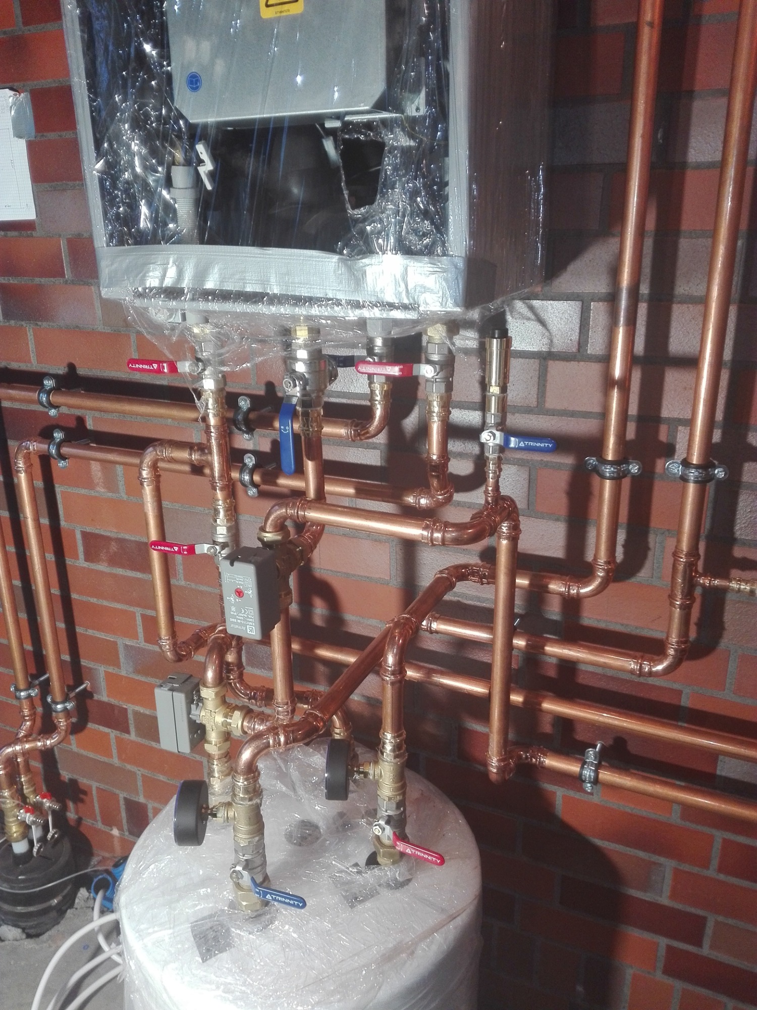 Nowo zainstalowany, miedziany system rur przyłączeniowych do pieca grzewczego i zasobnika wody, częściowo owinięty folią ochronną, na tle ściany z czerwonej cegły.