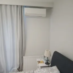 Klimatyzator ścienny zamontowany w sypialni nad grzejnikiem, widoczny fragment ł&oacute;żka z szarą pościelą w kwiatowy wz&oacute;r, zasłonięte okno i szafka nocna z lampką.