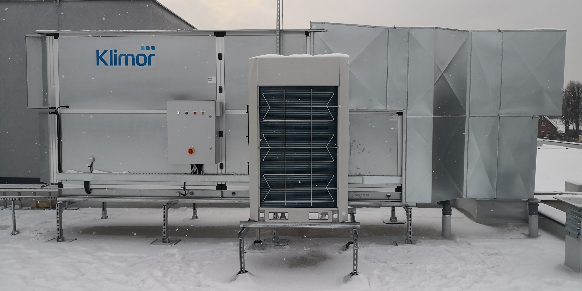 Instalacja wentylacyjna marki Klimor na dachu budynku, widoczny panel sterowania i kratka wentylacyjna, otoczenie pokryte śniegiem.