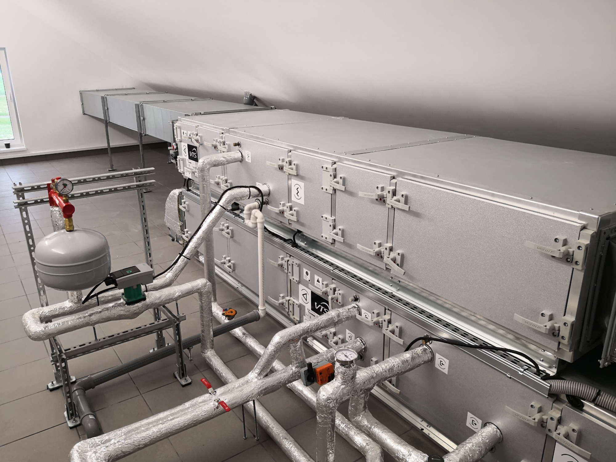 Instalacja rekuperacyjna w budynku komercyjnym: widok na długi, srebrny modułowy system kanałów wentylacyjnych pod sufitem, z licznymi rurami owiniętymi folią aluminiową, zaworami, manometrami...