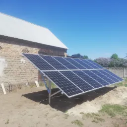 Panel słoneczny ustawiony na metalowej konstrukcji, na piaszczystym podłożu przed budynkiem gospodarczym z cegły i białym dachem, błękitne niebo w tle.