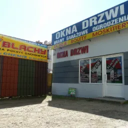 Fasada budynku firmy oferującej okna, drzwi, bramy garażowe, rolety i moskitiery, z widocznymi szyldami reklamowymi i ekspozycją dachówki obok.