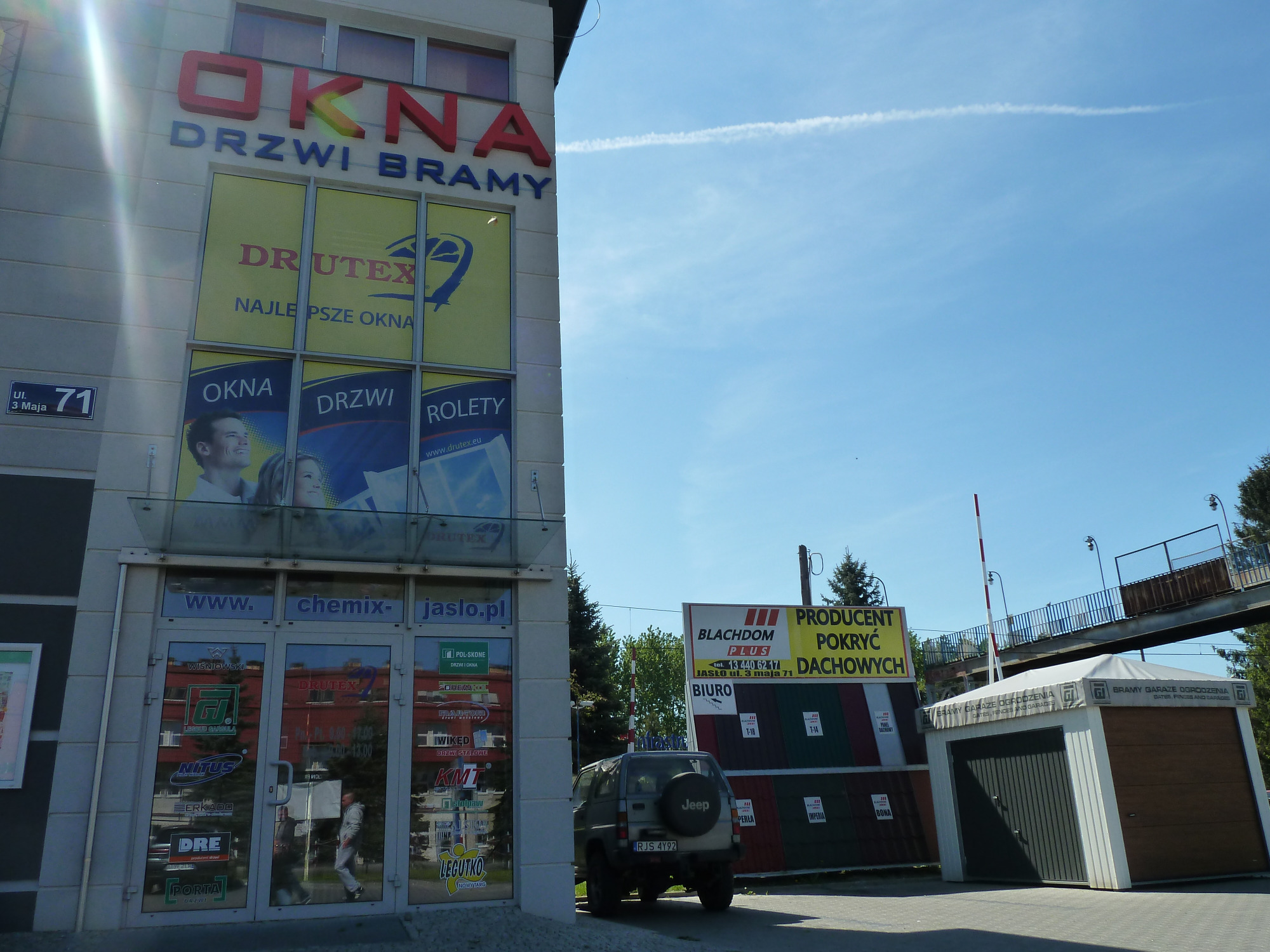 Fasada budynku firmy sprzedającej okna, drzwi i bramy w słoneczny dzień, z reklamami producentów i ekspozycją produktów w witrynach.