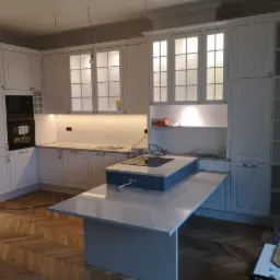 Jasna kuchnia z białymi szafkami, wyspą z szarym blatem i parkietem ułożonym we wzór jodełki; widoczne niedokończone prace elektryczne.