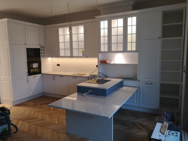 Jasna kuchnia z białymi szafkami, wyspą z szarym blatem i parkietem ułożonym we wzór jodełki; widoczne niedokończone prace elektryczne.