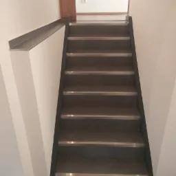 Wykończone schody z ciemnych płytek z metalowymi listwami antypoślizgowymi, białe ściany, widok z góry na dół.