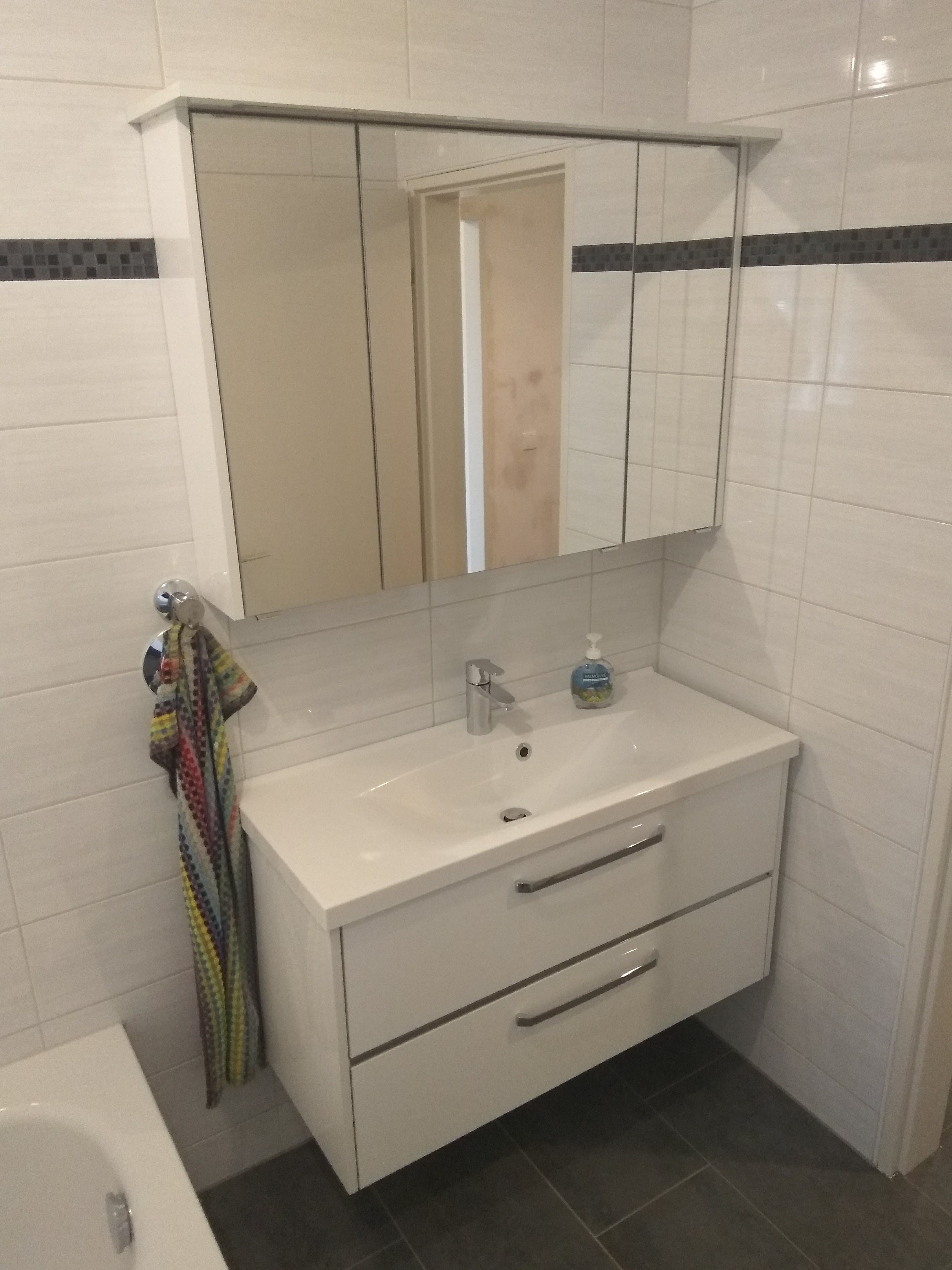 Nowoczesna łazienka z białą umywalką z szafką i lustrem w szafce, ręcznik w kolorowe paski po lewej stronie, płytki imitujące drewno na ścianach i szare płytki podłogowe.