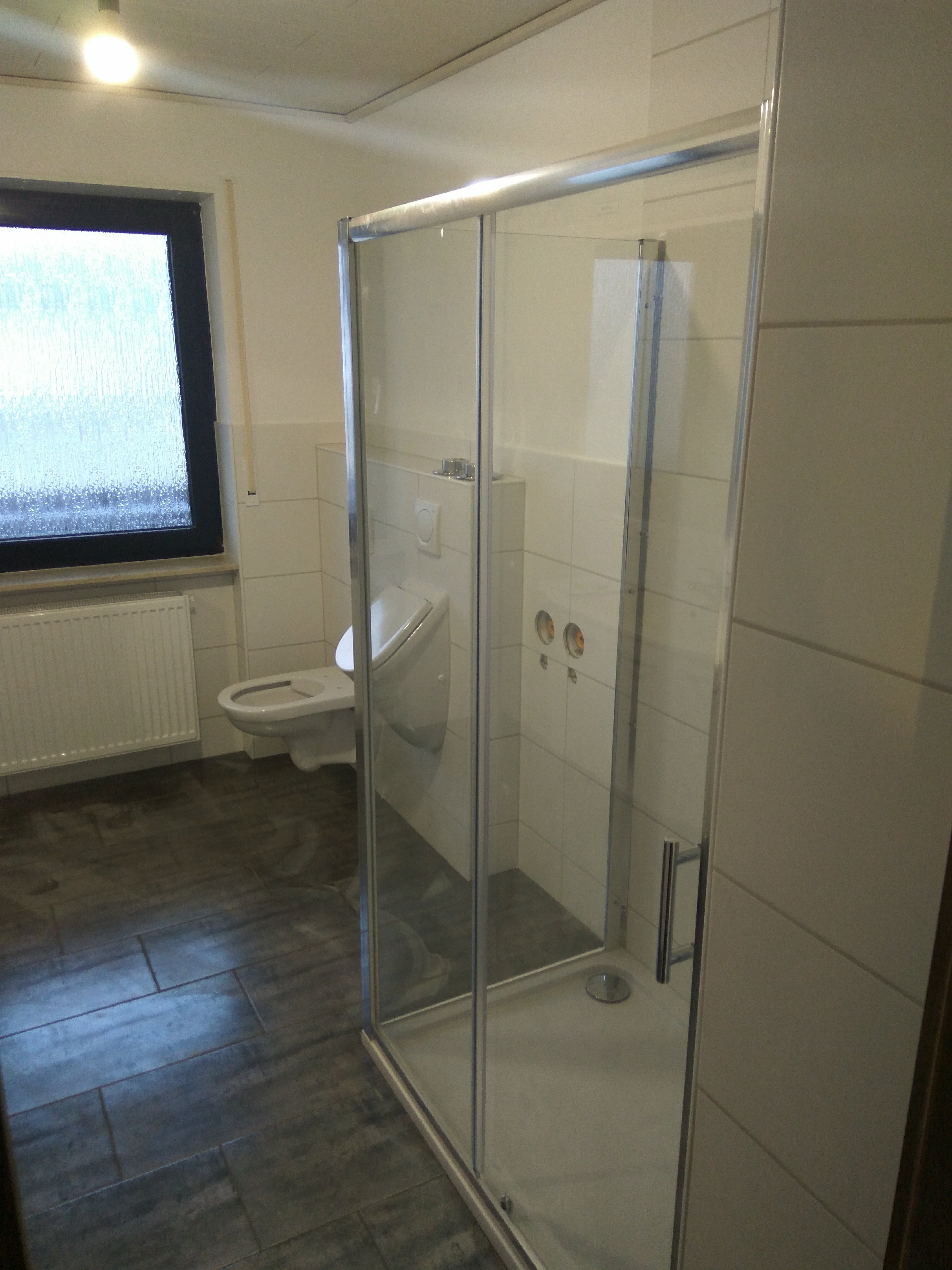 Nowoczesna kabina prysznicowa z przesuwnymi drzwiami ze szkła hartowanego, widoczny fragment toalety i grzejnik na tle ciemnej, płytkowej podłogi w jasnej łazience.