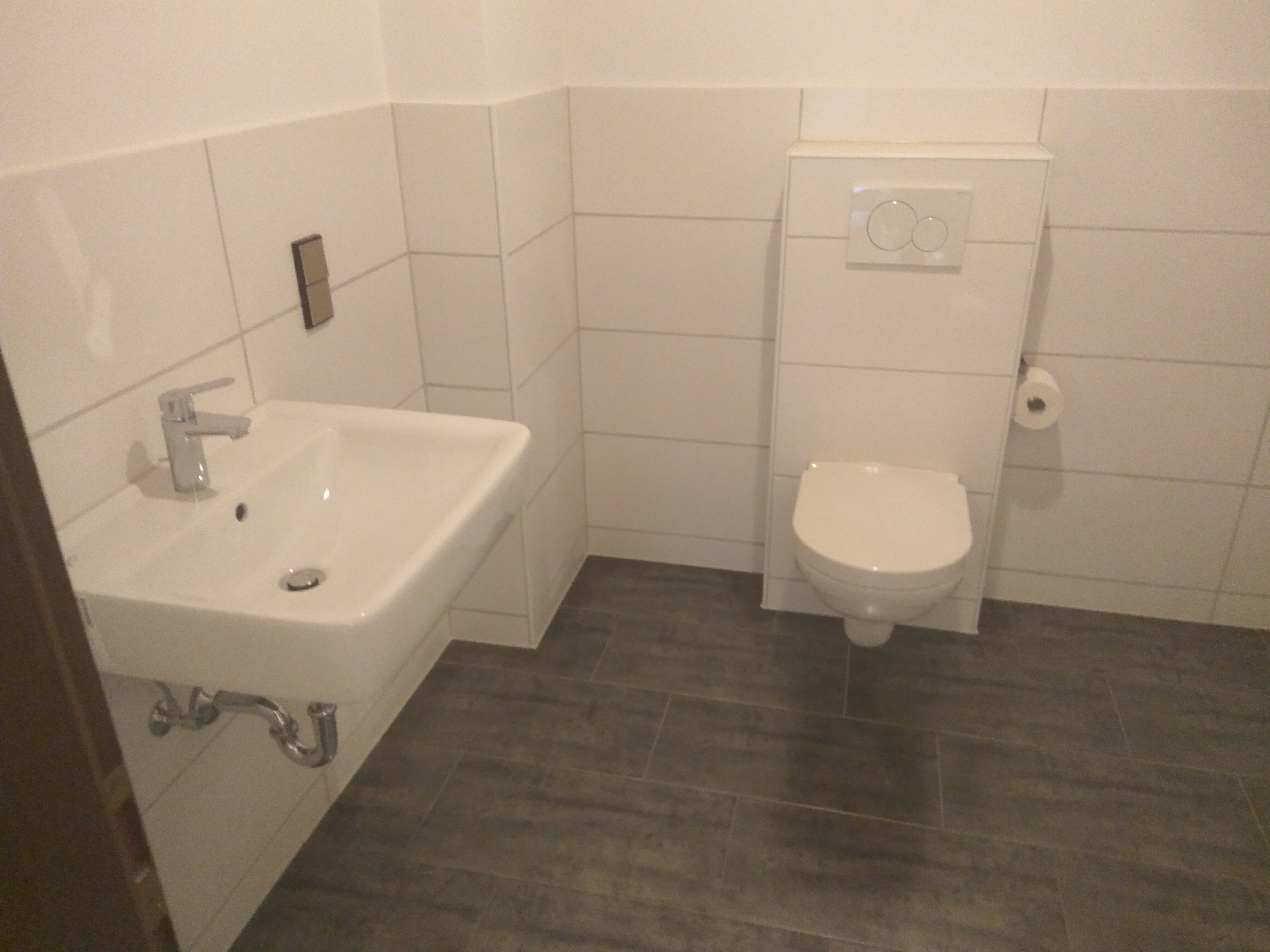 Biała umywalka wisząca z chromowaną baterią i odpływem, zamontowana na ścianie wyłożonej białymi płytkami; w tle podwieszana toaleta z białym przyciskiem spłukującym i rolką papieru toaletowego,...