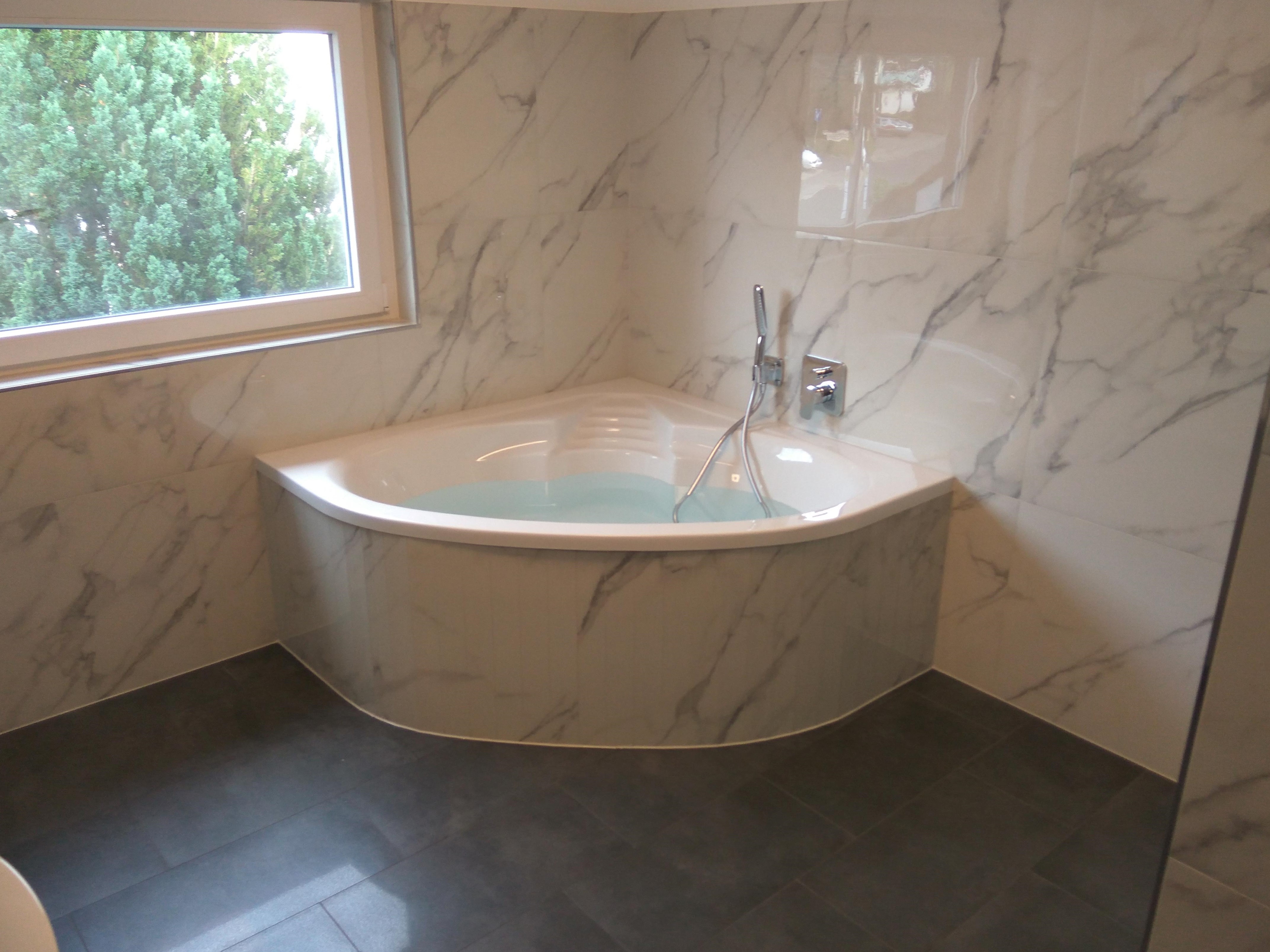 Narożna wanna wypełniona wodą, obudowana płytkami imitującymi marmur, z widoczną armaturą i oknem z zielenią w tle, na podłodze ciemne płytki.