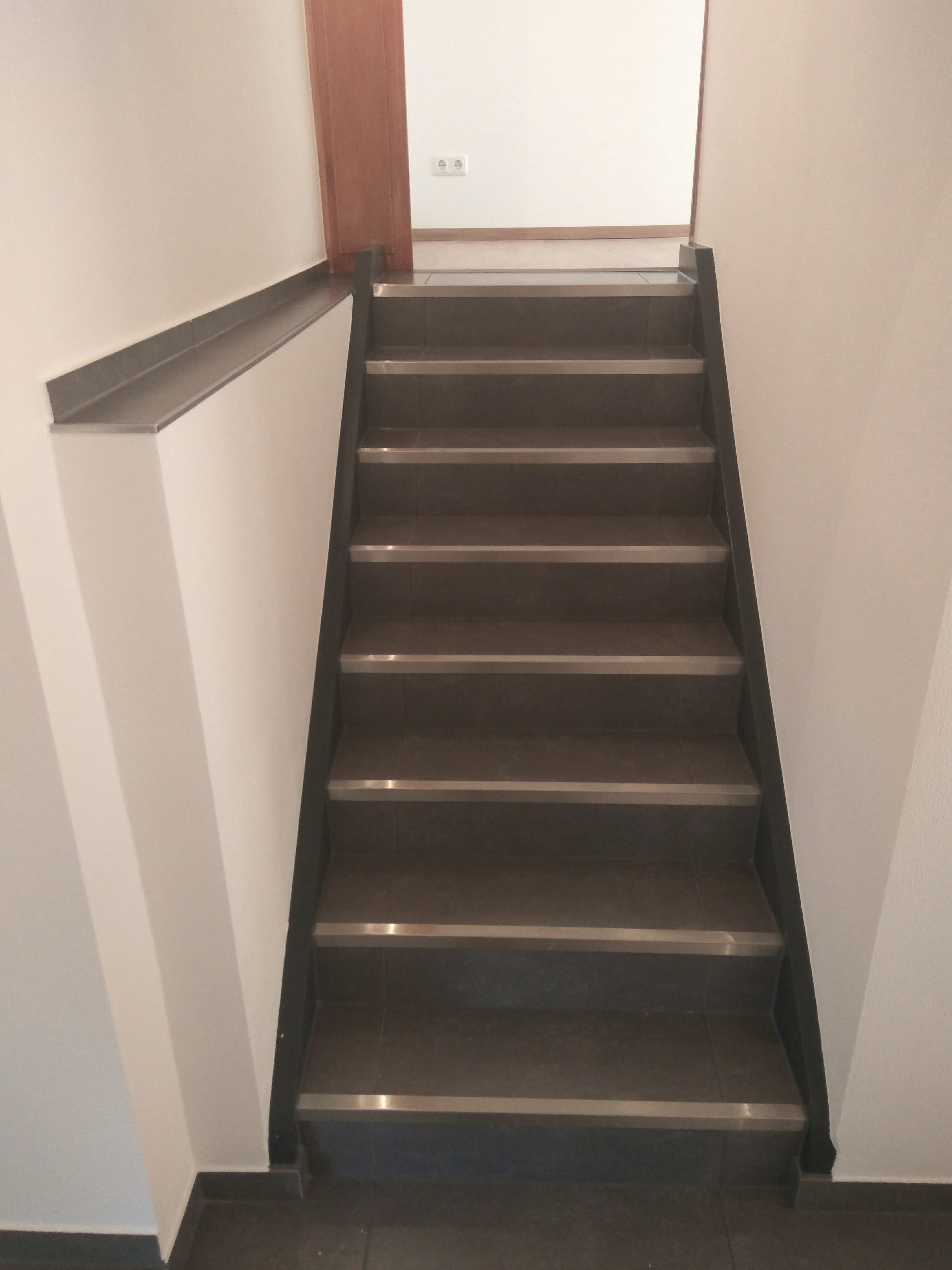 Wykończone schody z ciemnych płytek z metalowymi listwami antypoślizgowymi, białe ściany, widok z góry na dół.