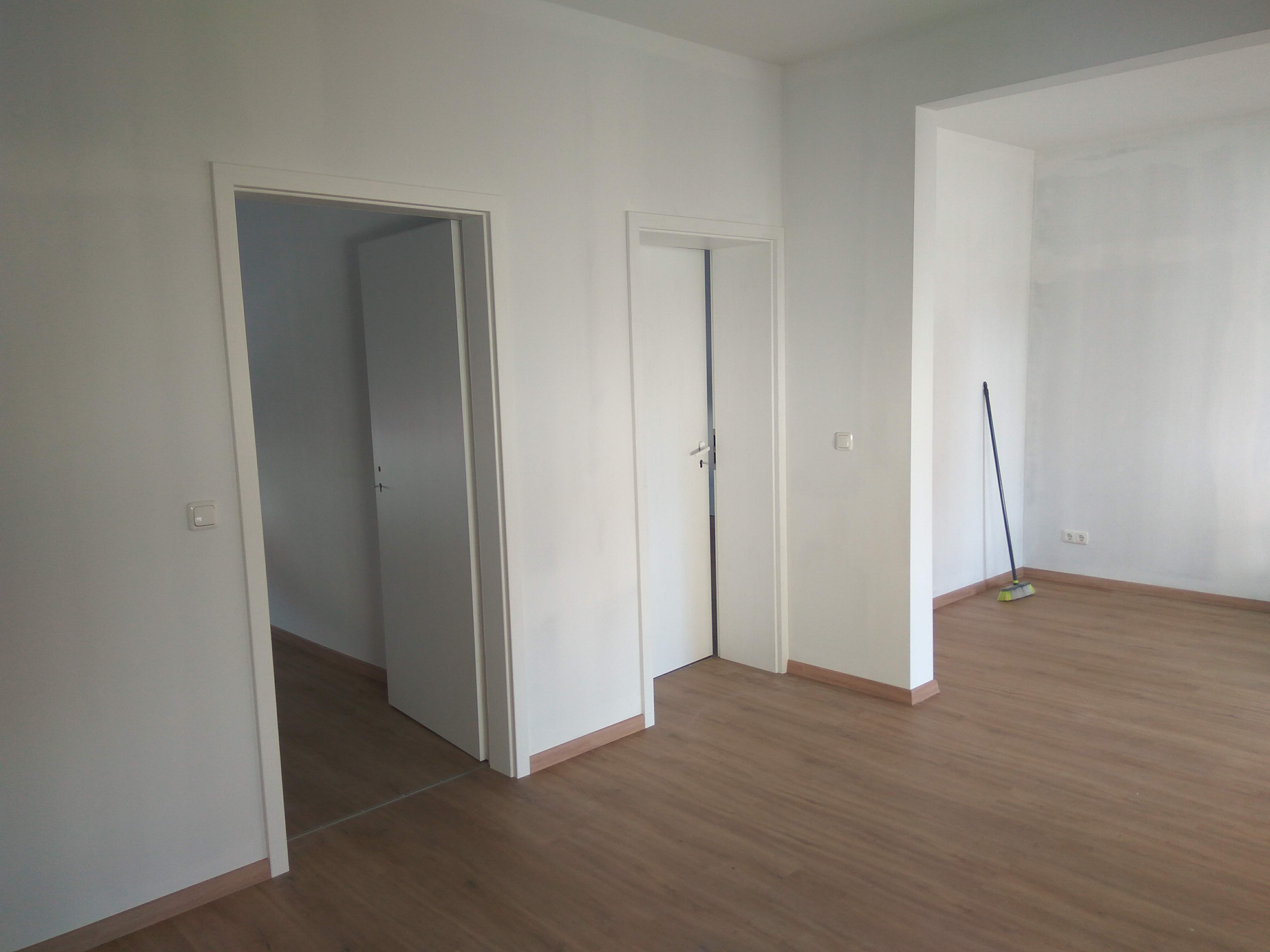 Pomalowane na biało wnętrze mieszkania w Aleksandrowie z dwiema ościeżnicami i drzwiami oraz panelami podłogowymi, w rogu stoi miotła.