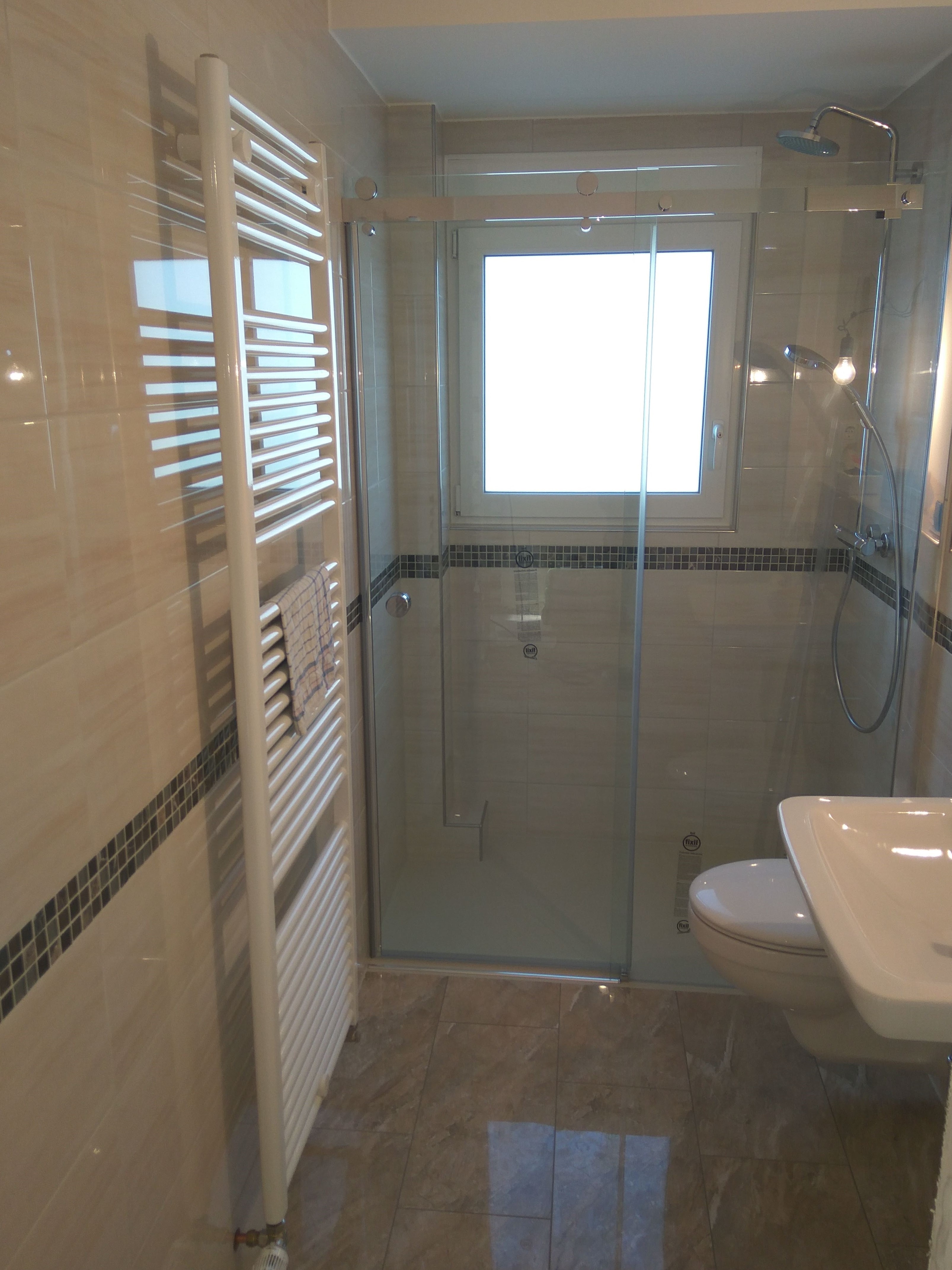 Nowoczesna łazienka z kabiną prysznicową ze szklanymi drzwiami przesuwnymi, grzejnikiem drabinkowym z zawieszonym ręcznikiem i umywalką z toaletą w tle, wykończona beżowymi płytkami z dekoracyjnym...